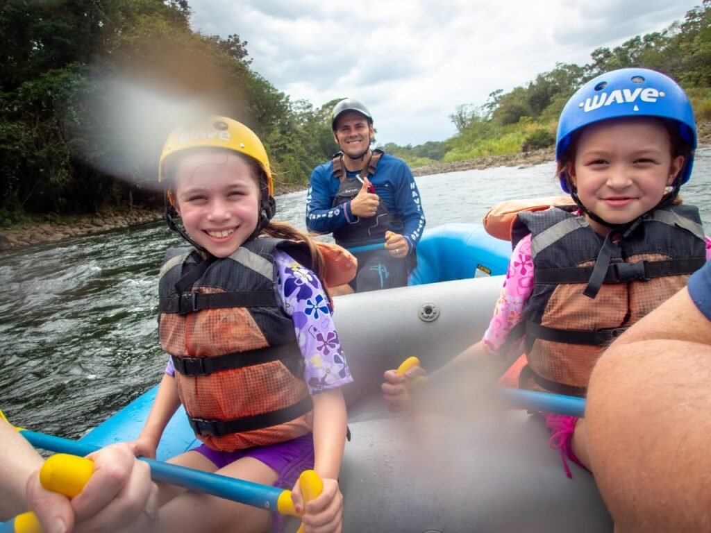 A group wearing helmets and life jackets enjoys rafting on a river, with smiles and water spray visible.