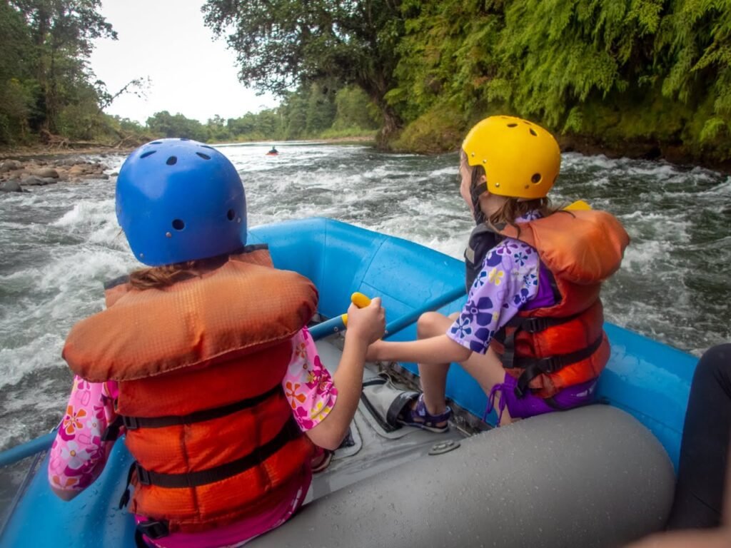 Two individuals wearing helmets and life jackets in a blue raft navigating fast-moving river waters.
