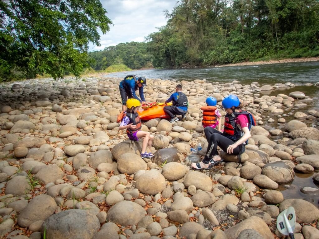 A group of people in helmets and life jackets sit on large river rocks next to a calm, forested river with a yellow raft in a tropical environment.
