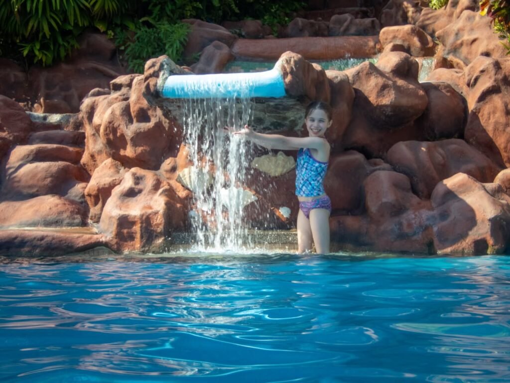 A child stands in a bright blue pool water beneath a waterfall feature surrounded by large rocks.
