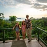 A family enjoys a ride on a swing overlooking a lush tropical forest and the ocean from a high vantage point.