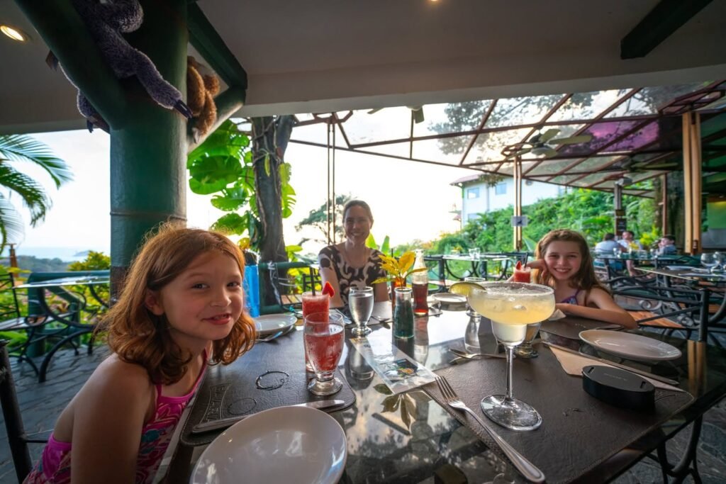 A family enjoys beverages at an outdoor tropical restaurant with a monkey on a beam above.