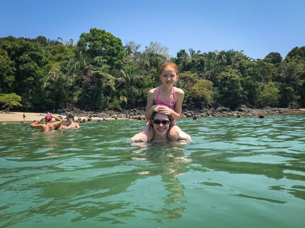 Two people enjoy a beautiful, clear body of water surrounded by lush tropical foliage and a rocky shoreline. A separate view shows a person in a pink top against a backdrop of dense, green trees under a blue sky.
