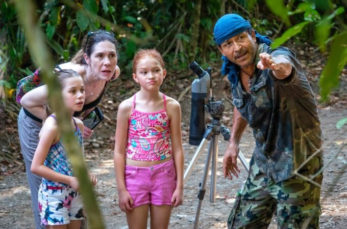 A group of people in a lush jungle, featuring a guide pointing forward and a tripod with a telescope set up nearby.
