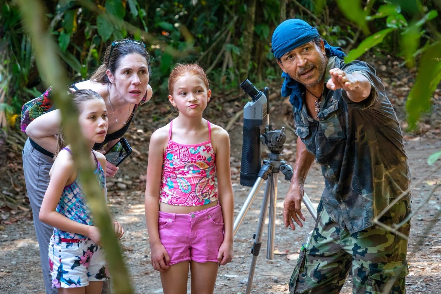 A group of people in a lush jungle, featuring a guide pointing forward and a tripod with a telescope set up nearby.