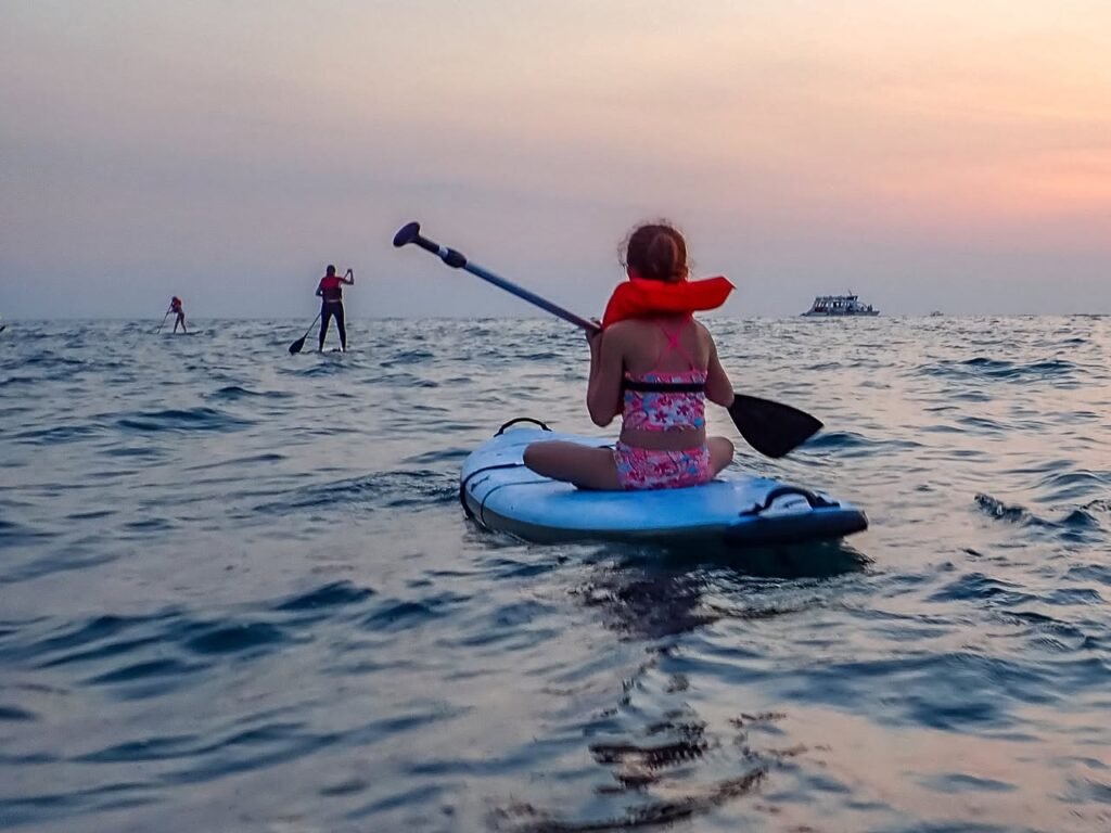 A person paddling a blue board at sunset with calm ocean water. Other paddleboarders are visible in the distance under a soft pink sky.