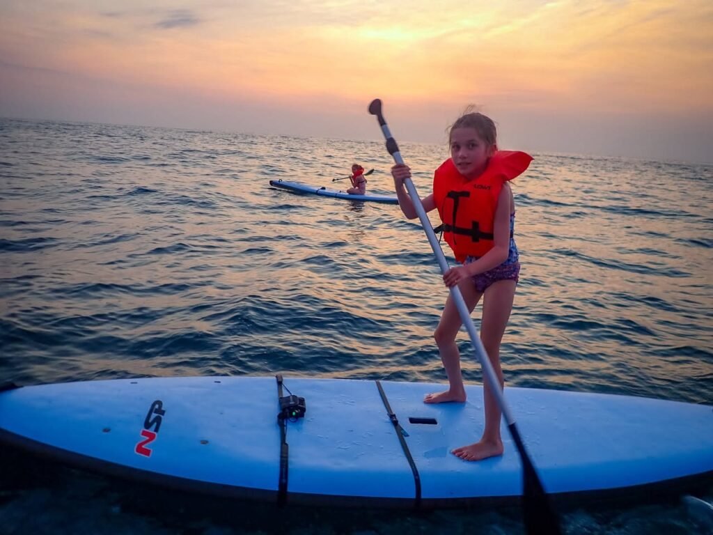 A child stands on a paddle board wearing a life jacket during a beautiful sunset, with calm ocean waters and warm skies.
