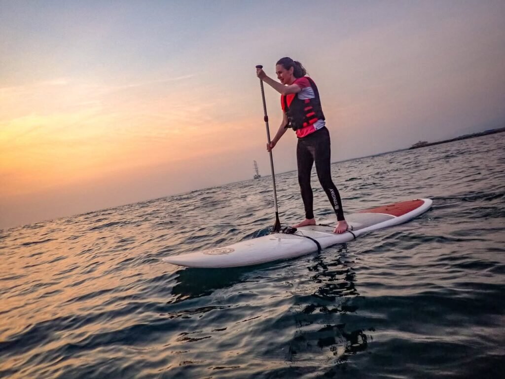 A person standup paddleboarding on calm water at sunset with a distant sailboat visible on the horizon.