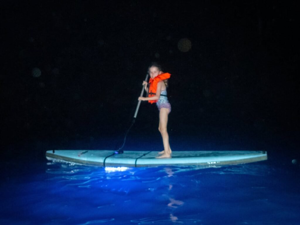 A person in an orange life vest paddleboards at night on water illuminated by blue lights. The dark background contrasts with the glowing water and illuminated figure.