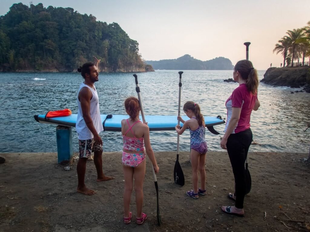 A family stands by a paddleboard and paddles on a waterfront concrete bank. The background features a sunset over water and tropical hills.