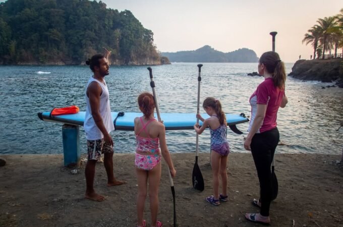 A family stands by a paddleboard and paddles on a waterfront concrete bank. The background features a sunset over water and tropical hills.