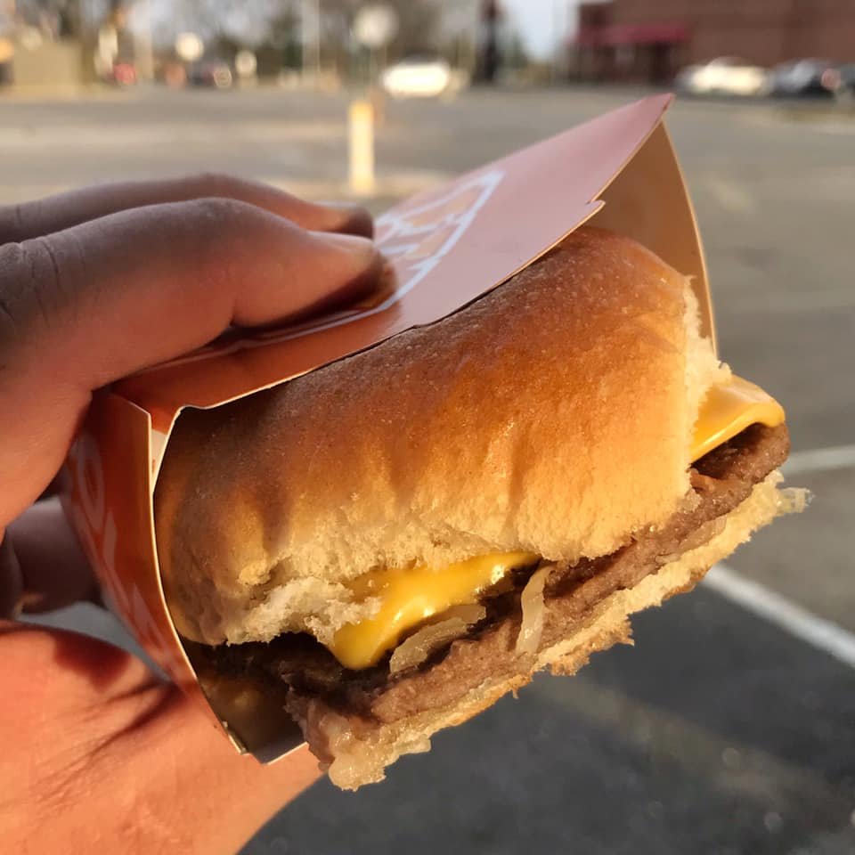 A hand holds a cheeseburger wrapped in paper outdoors with a cloudy background.