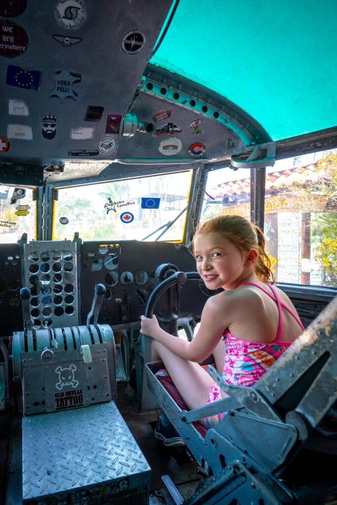 A child sits in the driver's seat of an excavator cab looking at the camera.