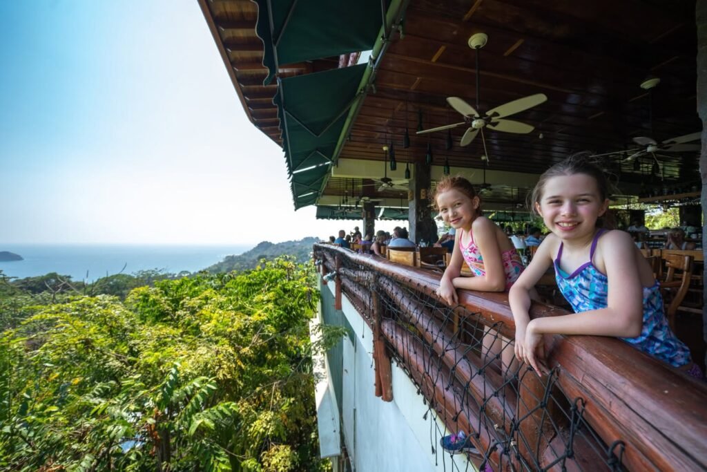 People leaning on a wooden balcony railing overlooking lush green treetops and distant ocean under a bright sky.