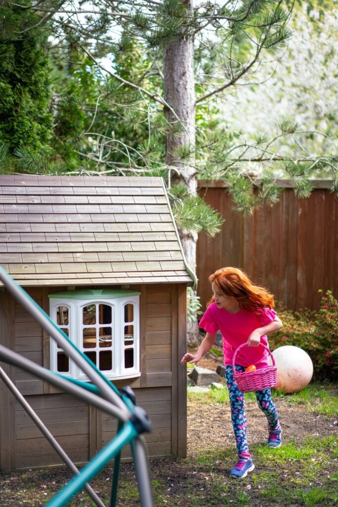 A young girl holding a pink basket walks near a basketball hoop and a backyard playhouse while searching for Easter eggs.