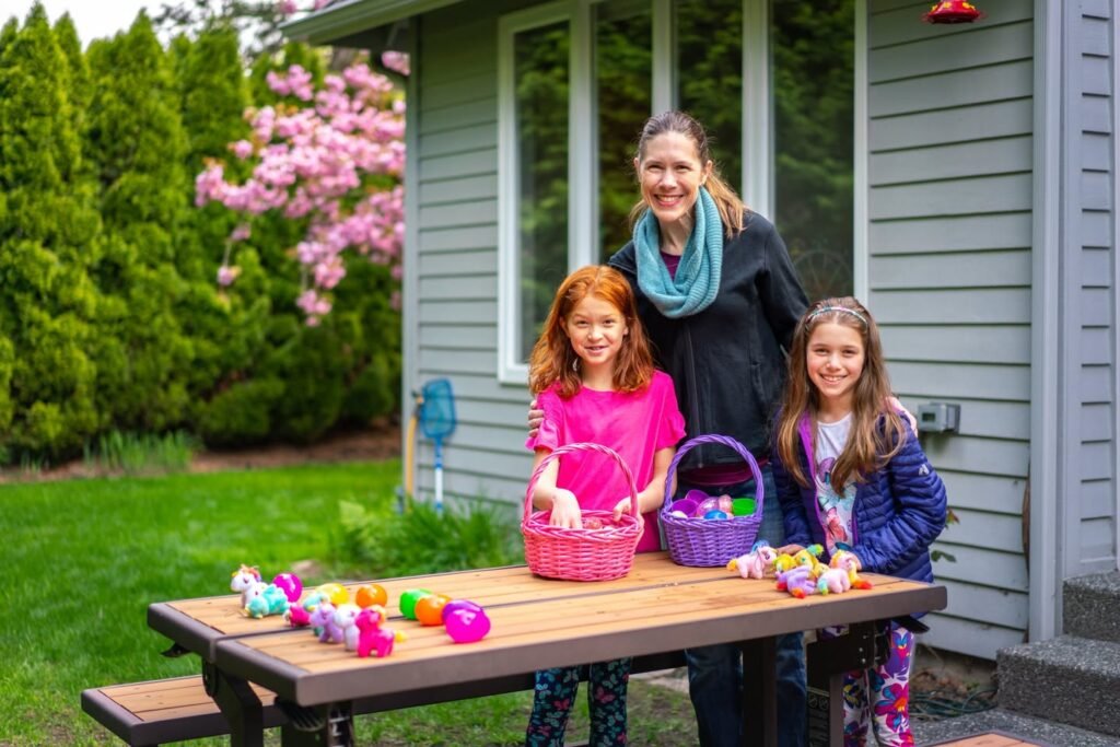 Family members with Easter egg baskets at a wooden picnic table. Brightly colored eggs decorate the table in a sunny garden.