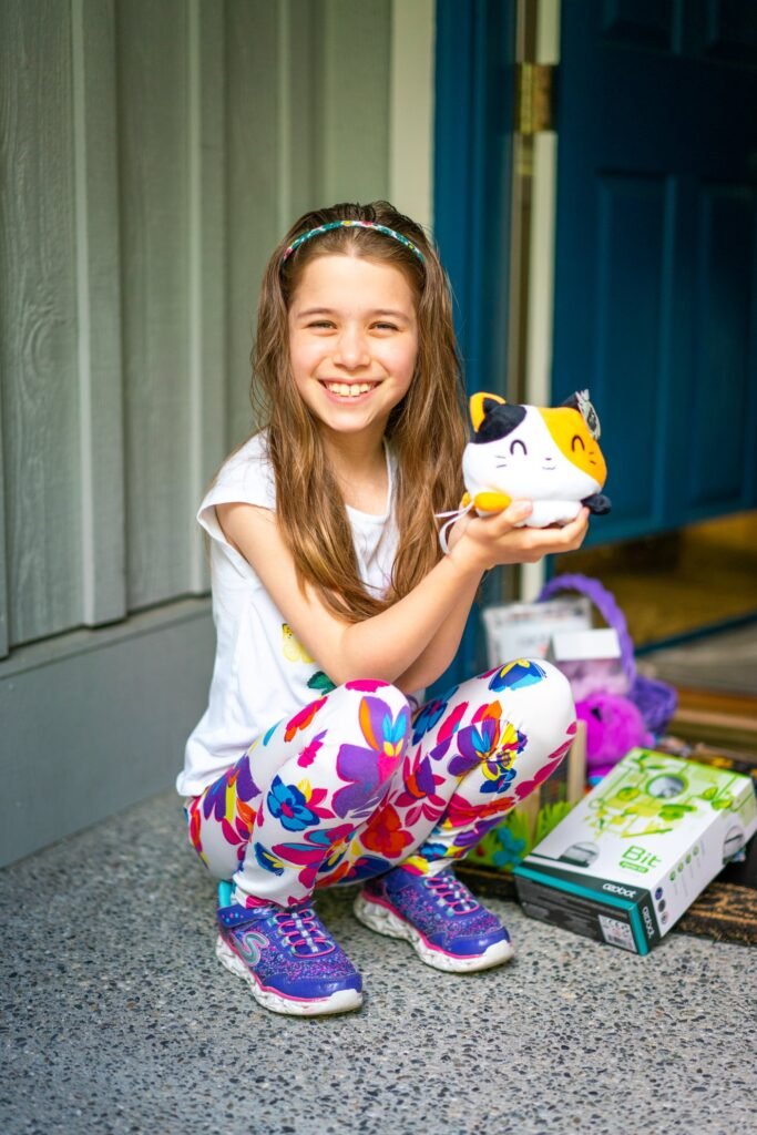 A smiling young person crouches on a porch holding a plush toy cat, beside a gift basket with a book.