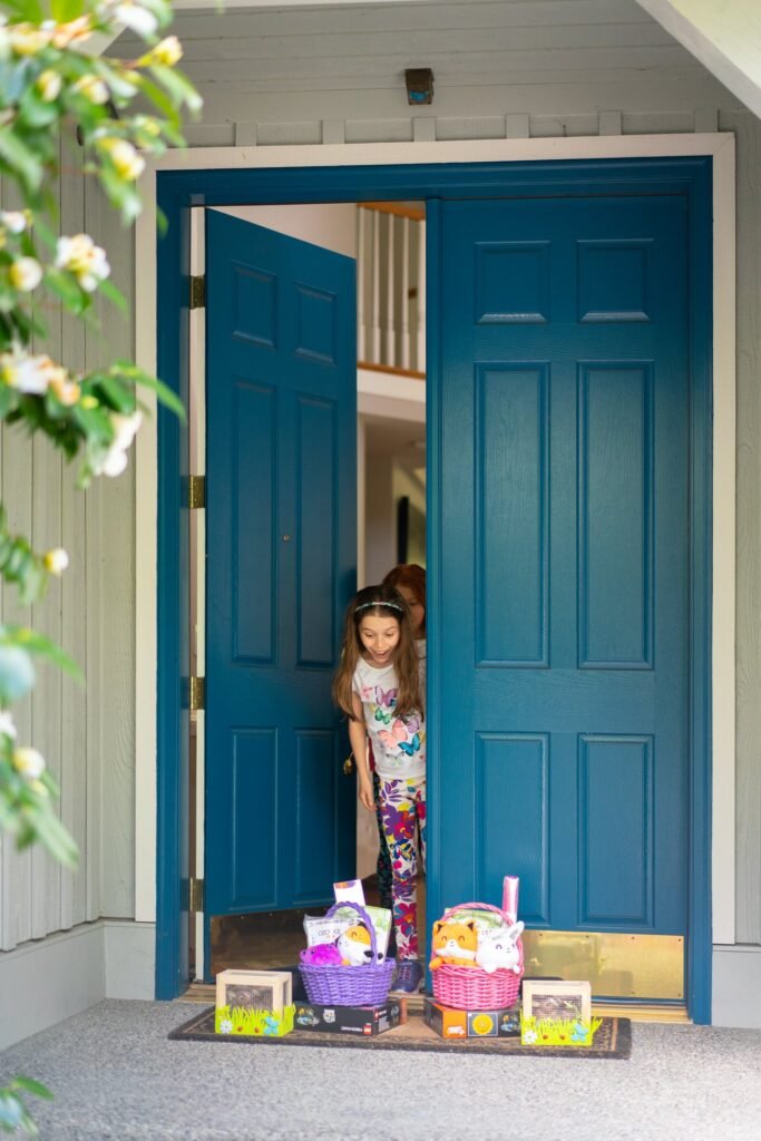 A child stands in a teal front doorway with Easter baskets and eggs at their feet.