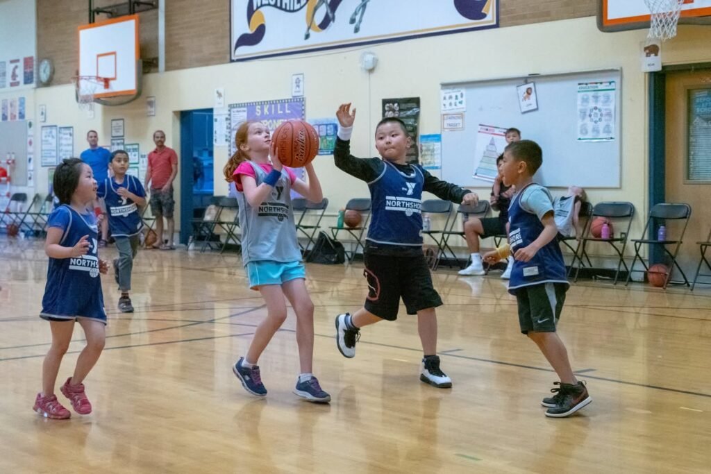 Young players competing in a basketball game in an indoor gymnasium. Spectators watch from the sidelines during the match.