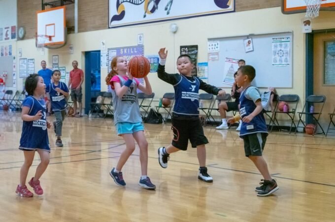 Young players competing in a basketball game in an indoor gymnasium. Spectators watch from the sidelines during the match.