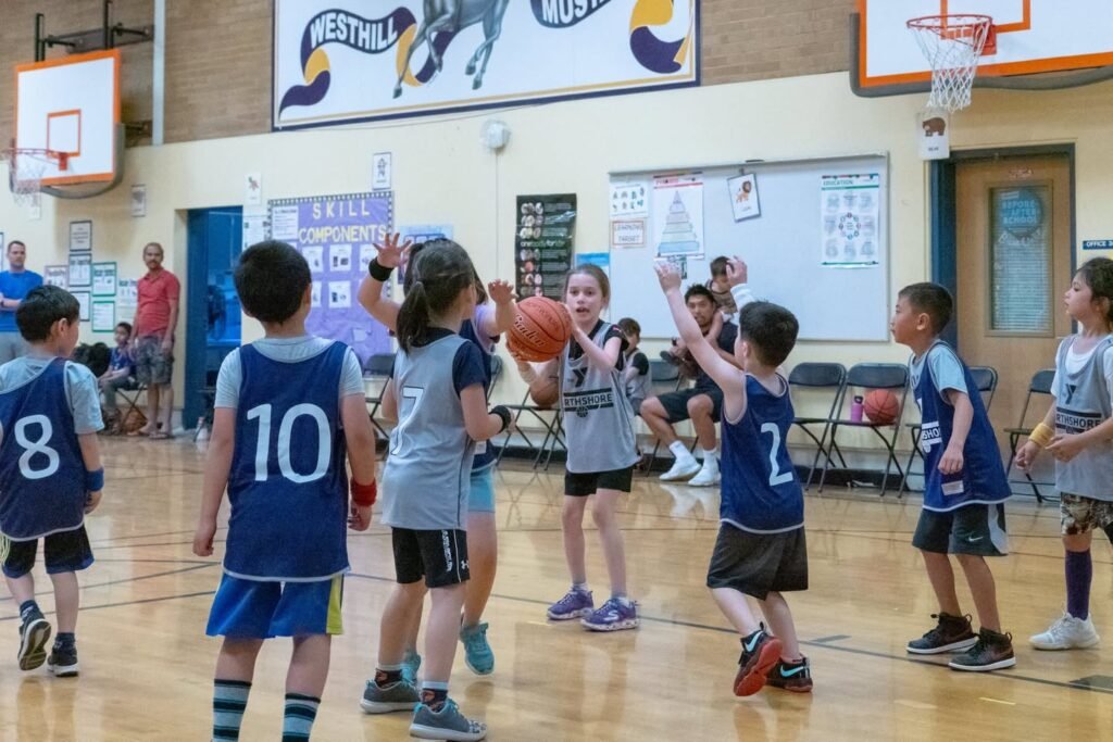 Young children playing an indoor basketball game in a school gymnasium, with a focus on the court and players in team jerseys.