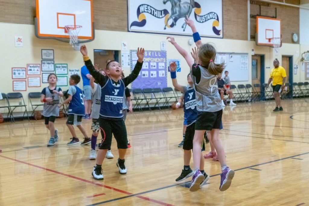 Young players leaping for the ball during an indoor basketball game. The gym features a wooden floor and school banners on the wall.