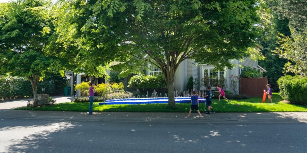 Several individuals wearing colorful swimwear play in a swimming pool surrounded by lush green trees and a white house in the backyard.