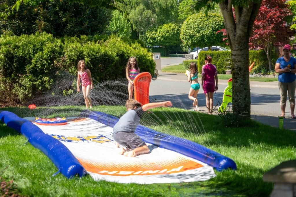 A child slides off an inflatable backyard water slide while family members watch and play with a garden hose.