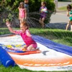 Children playing on an inflatable slip and slide in a backyard sprinkler setup.