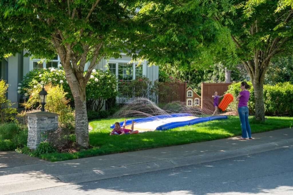 Children playing in a sprinkler on a sunny day. Lush green trees and a suburban street complete the scene.