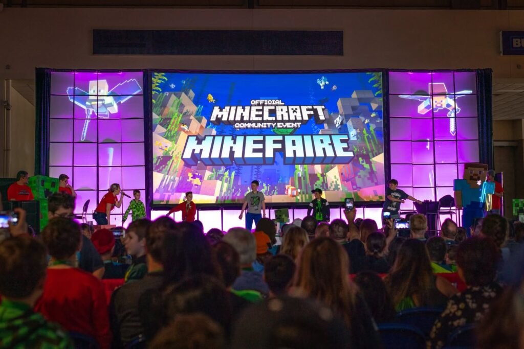 The team performs on stage at Minefaire with an audience watching them. A large screen behind them displays the Minecraft and Minefaire logos.