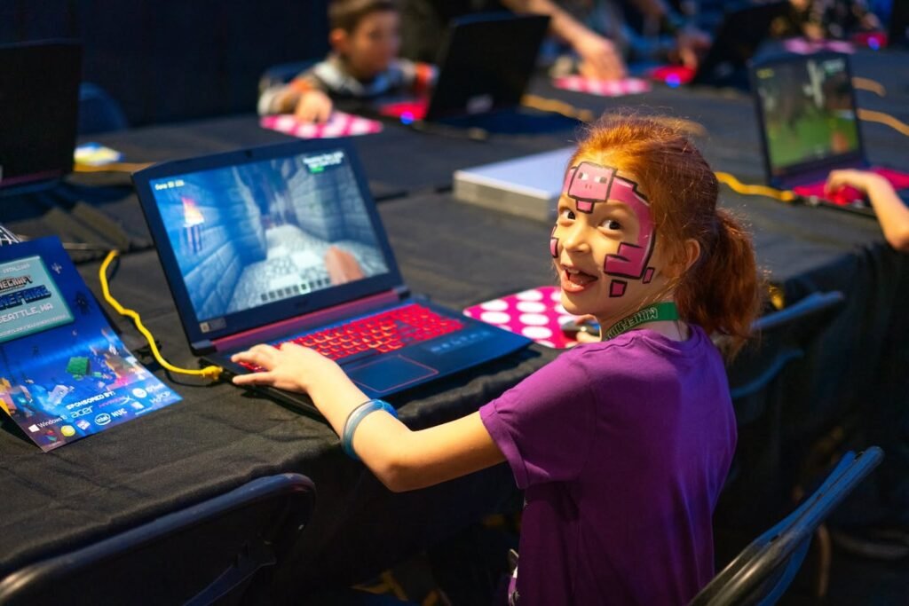 A child with a DMC face painted smiles while using a laptop at a young gamers competition. Other participants are also focused on their computers.