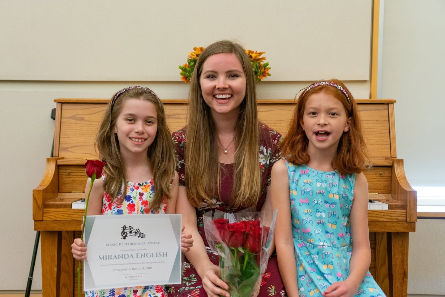 Spring 2019 Piano Recital Three individuals posing with a flower and certificate in front of a wooden upright piano.