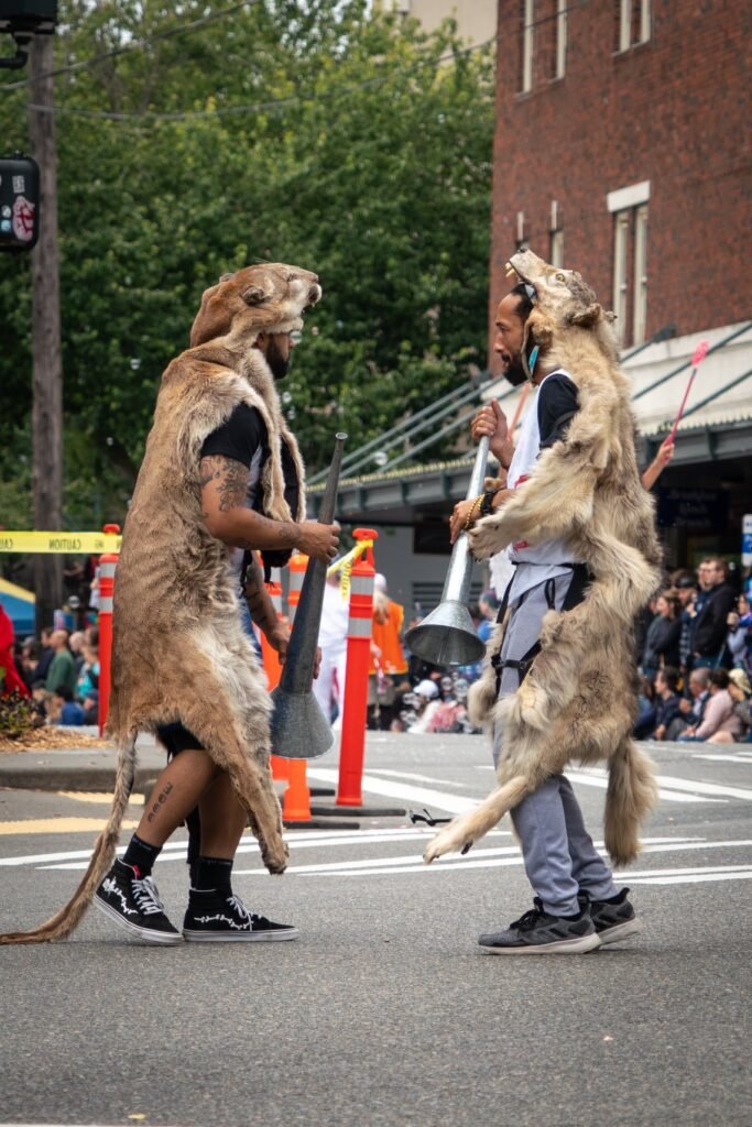 Festival participants wearing kangaroo costumes performing in a public parade. Two costumed musicians play instruments while marching down an urban street lined with cheering spectators.
