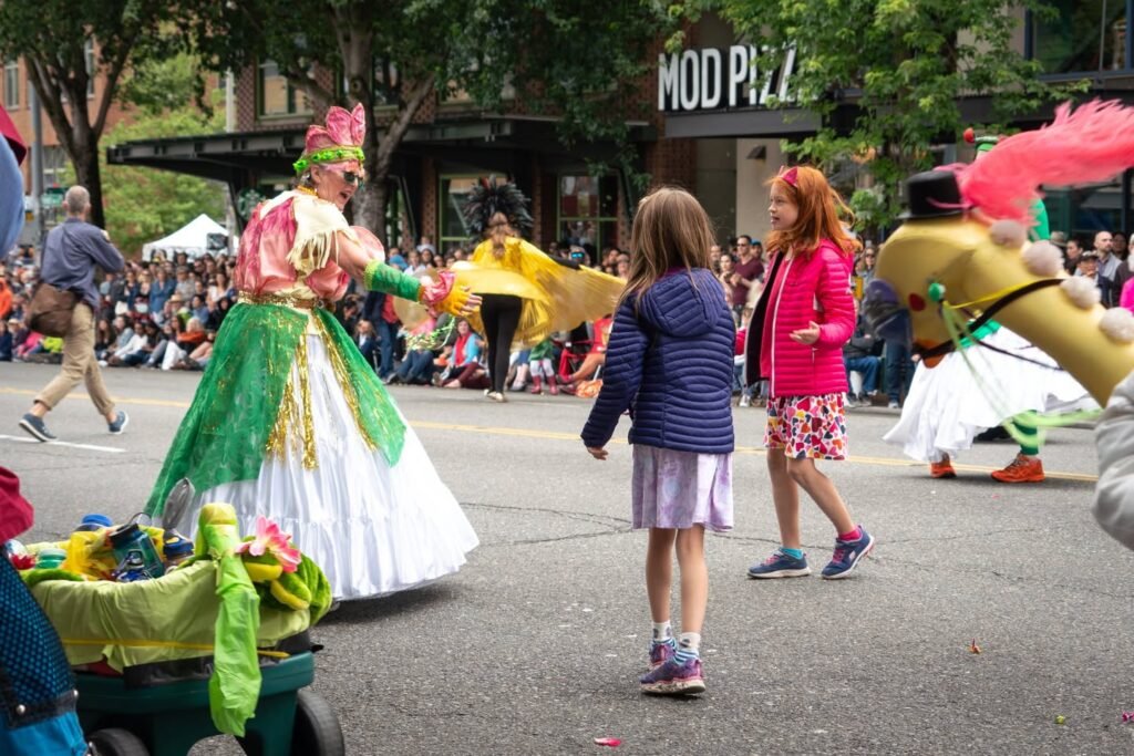 Colorful parade participants in elaborate costumes walk through a lively street festival. Spectators watch from the sidewalk as performers dance and interact with the crowd.