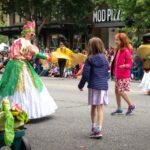 Cultus Lake, British Columbia Colorful parade participants in elaborate costumes walk through a lively street festival. Spectators watch from the sidewalk as performers dance and interact with the crowd.