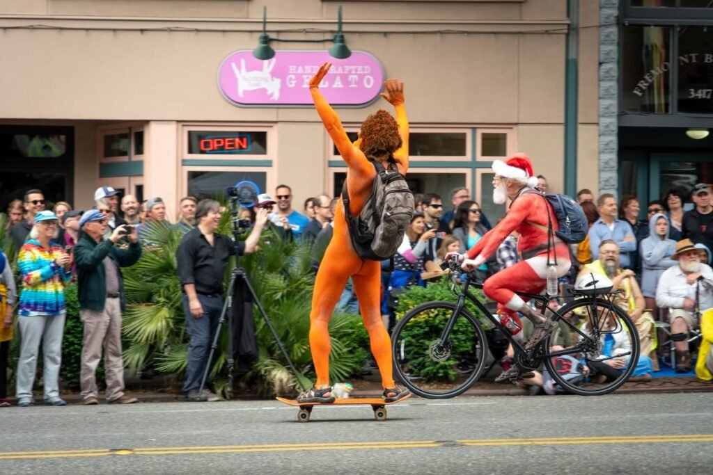 A person in orange skateboards and another in a Santa outfit rides a bike in front of a lively crowd. This festive scene captures the spirit of a street parade.