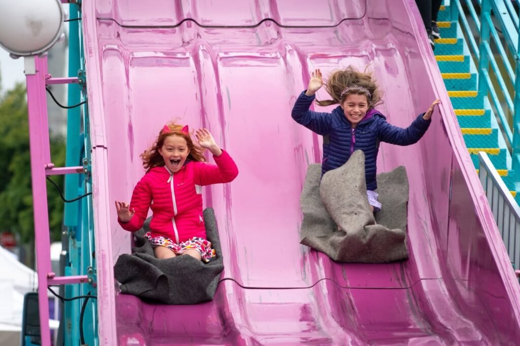 Two people riding a large pink playground slide. The structure features teal and yellow steps in the background.