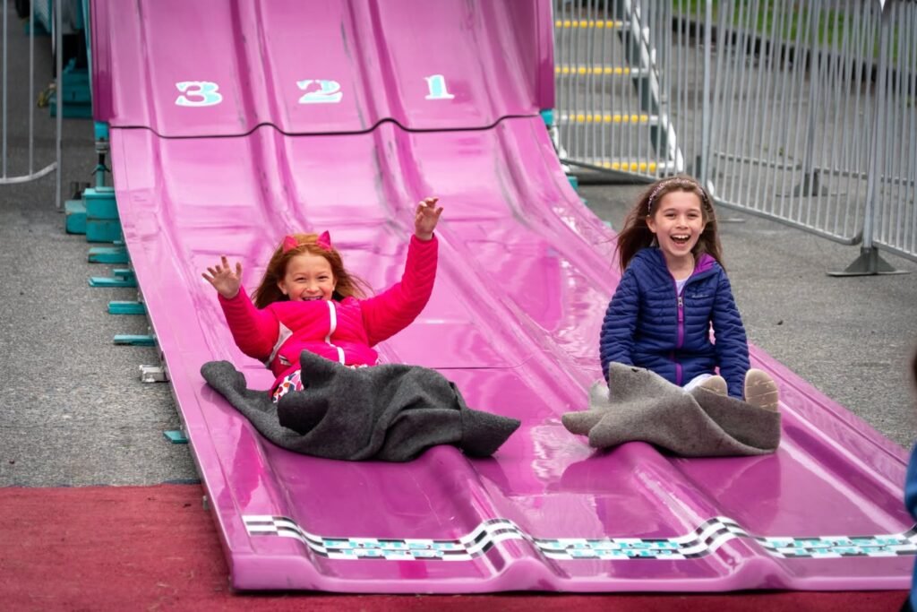 Two children sliding down a large pink inflatable chute with numbered lanes at an outdoor carnival.