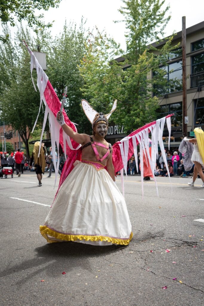 A person in a white dress with a yellow hem and metal headpiece waves a red and white banner on a city street during a parade, with crowds and buildings in the background.