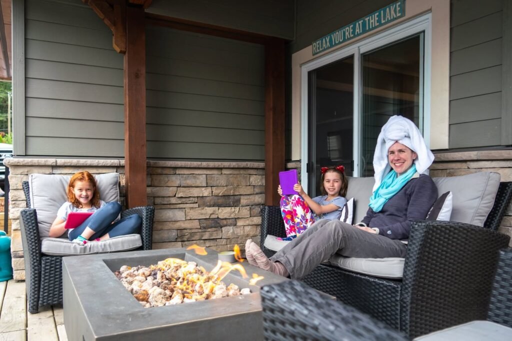 A family enjoying a cozy evening on a wooden deck with a gas fire pit. The setting includes an outdoor fireplace and stone wall at a lakefront lodge.