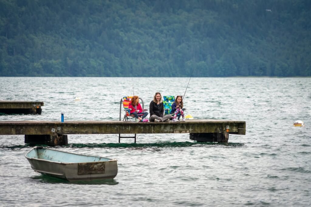 A family enjoys a day of fishing on a rustic dock by a serene lake. A small boat floats nearby under a misty mountain backdrop.