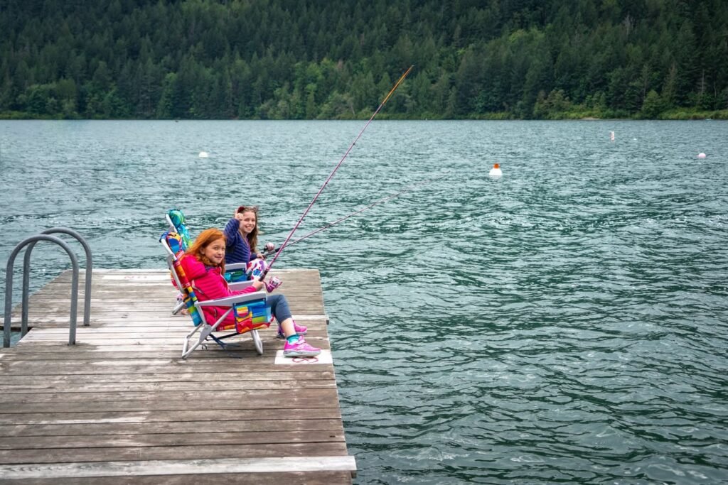 A relaxed scene of fishing from a wooden dock on a calm lake, surrounded by lush evergreen forest typical that of the Pacific Northwest.