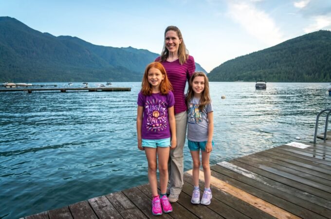 A woman and two girls standing together on a wooden dock in the Pacific Northwest. Lush mountains and a serene lake provide a beautiful scenic backdrop.
