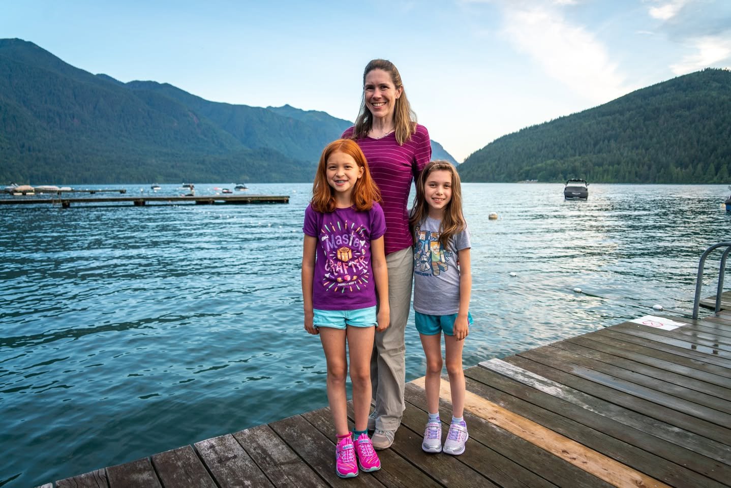 Cultus Lake, British Columbia A woman and two girls standing together on a wooden dock in the Pacific Northwest. Lush mountains and a serene lake provide a beautiful scenic backdrop.