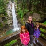 Cultus Lake, British Columbia A family of three smiles at a wooden viewpoint overlook. A tall waterfall cascades down a mossy rock face in the lush green background.