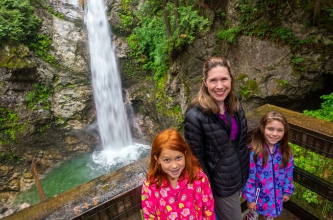 A family of three smiles at a wooden viewpoint overlook. A tall waterfall cascades down a mossy rock face in the lush green background.
