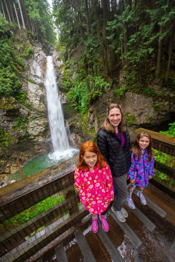 A family of three smiles at a wooden viewpoint overlook. A tall waterfall cascades down a mossy rock face in the lush green background.