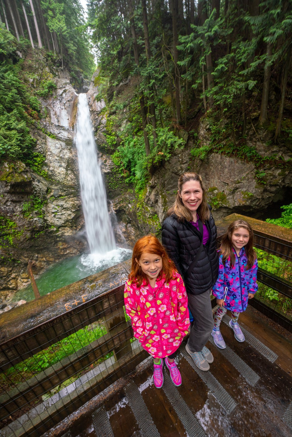 Cascade Falls A family of three smiles at a wooden viewpoint overlook. A tall waterfall cascades down a mossy rock face in the lush green background.