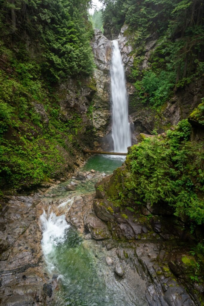 Waterfall cascading down a rugged cliff into a clear pool, surrounded by greenery. Parts of a rocky wall structure with patches of greenery on the right side.